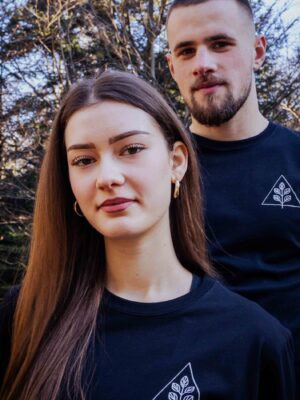 Modelpaar trägt schwarzes Unisex T-Shirt mit weißem Pflanzenmotiv und Dreieck