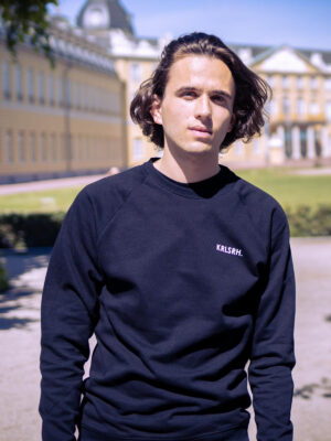Männliches Model trägt schwarzen Crewneck Pullover bedruckt mit KRLSRH. Schriftzug